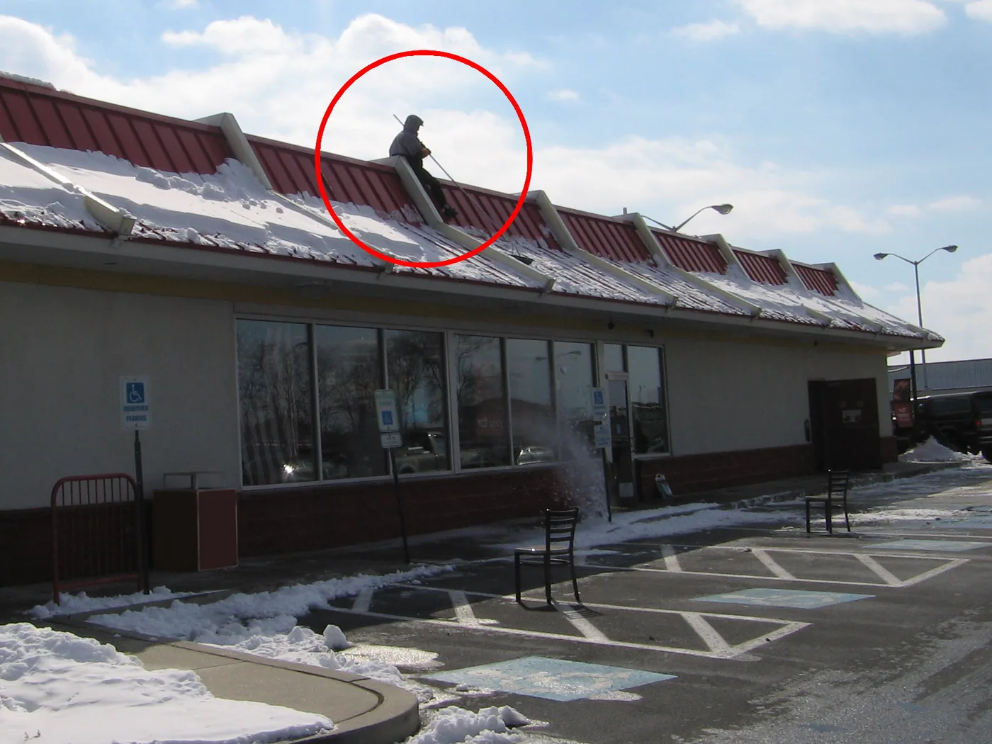 The sidewalk is closed off during business hours while an employee hangs off the side of the roof to manually remove snow from the panels